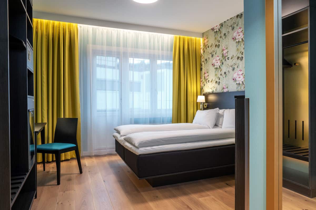 Una habitación de hotel moderna con una cama, una silla, un armario y una ventana con cortinas amarillas.