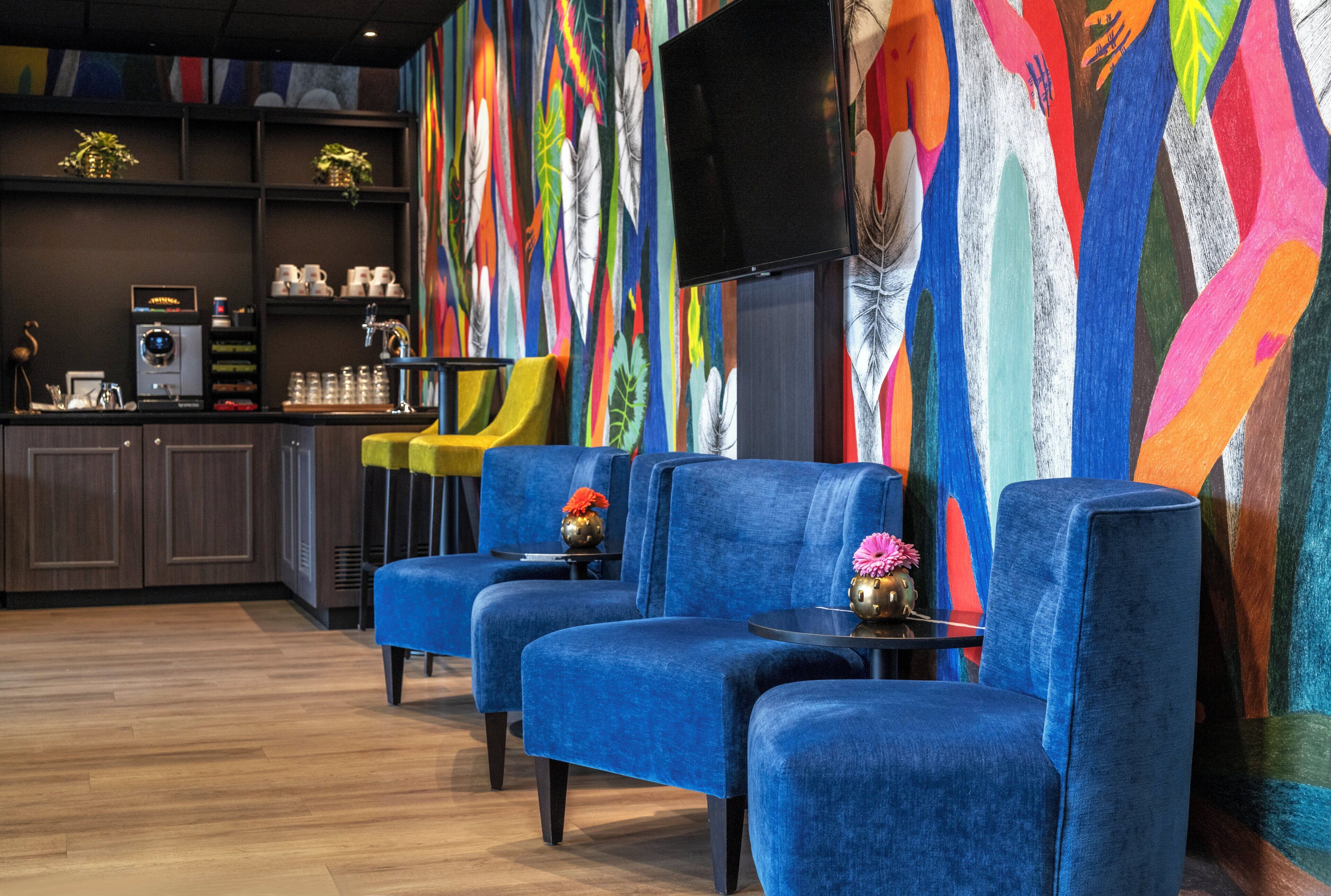 A modern lounge area with blue sofas, a coffee machine, and a colorful abstract wall.