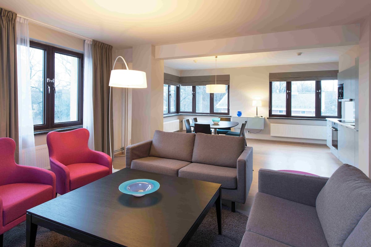 Habitación de hotel con zona de estar que incluye un sofá y dos sillones, una mesa de comedor con sillas, y ventanas amplias que dejan entrar luz natural.
