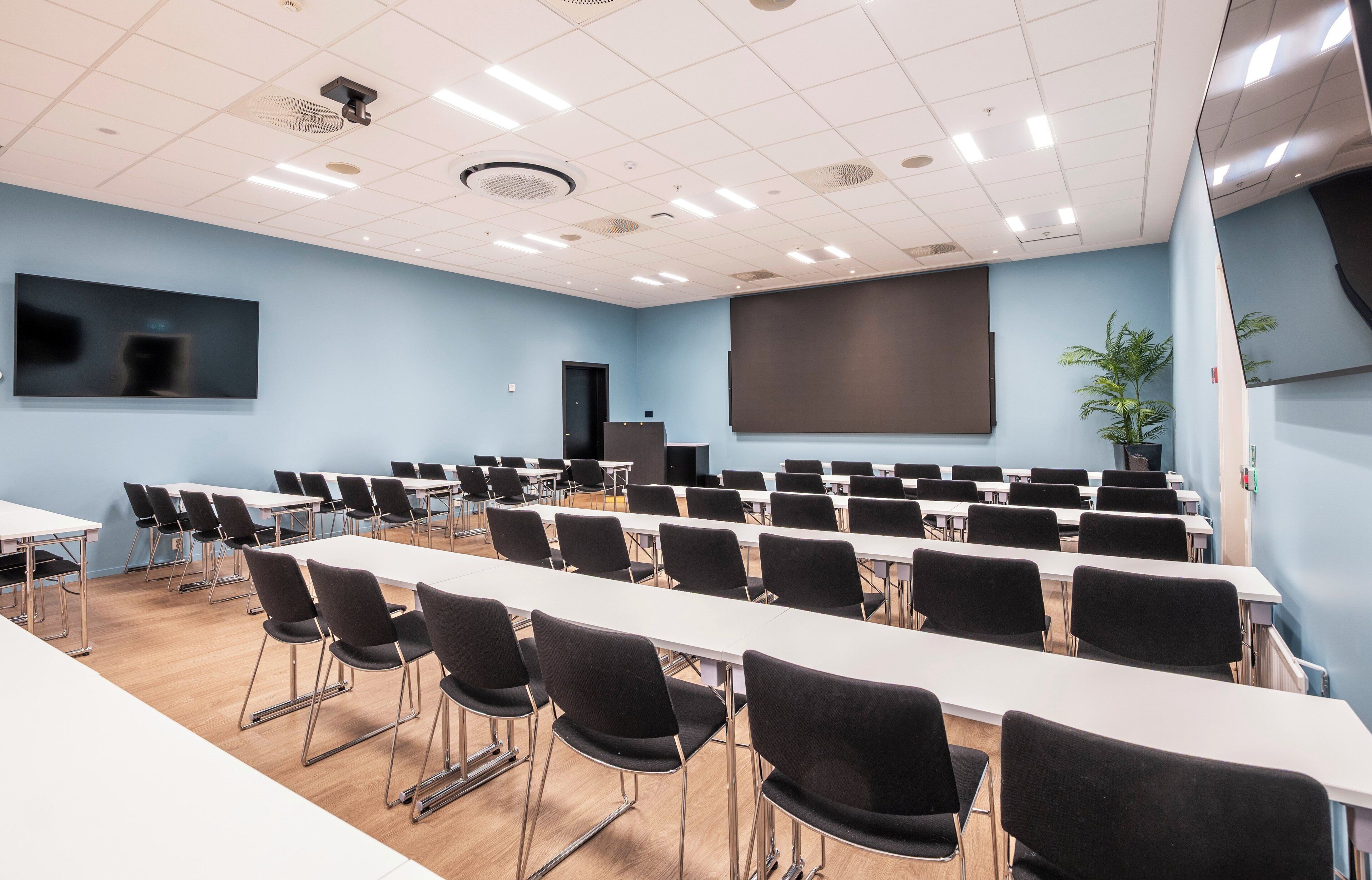 A conference room with tables and chairs, a projector screen, and a large screen on the wall.