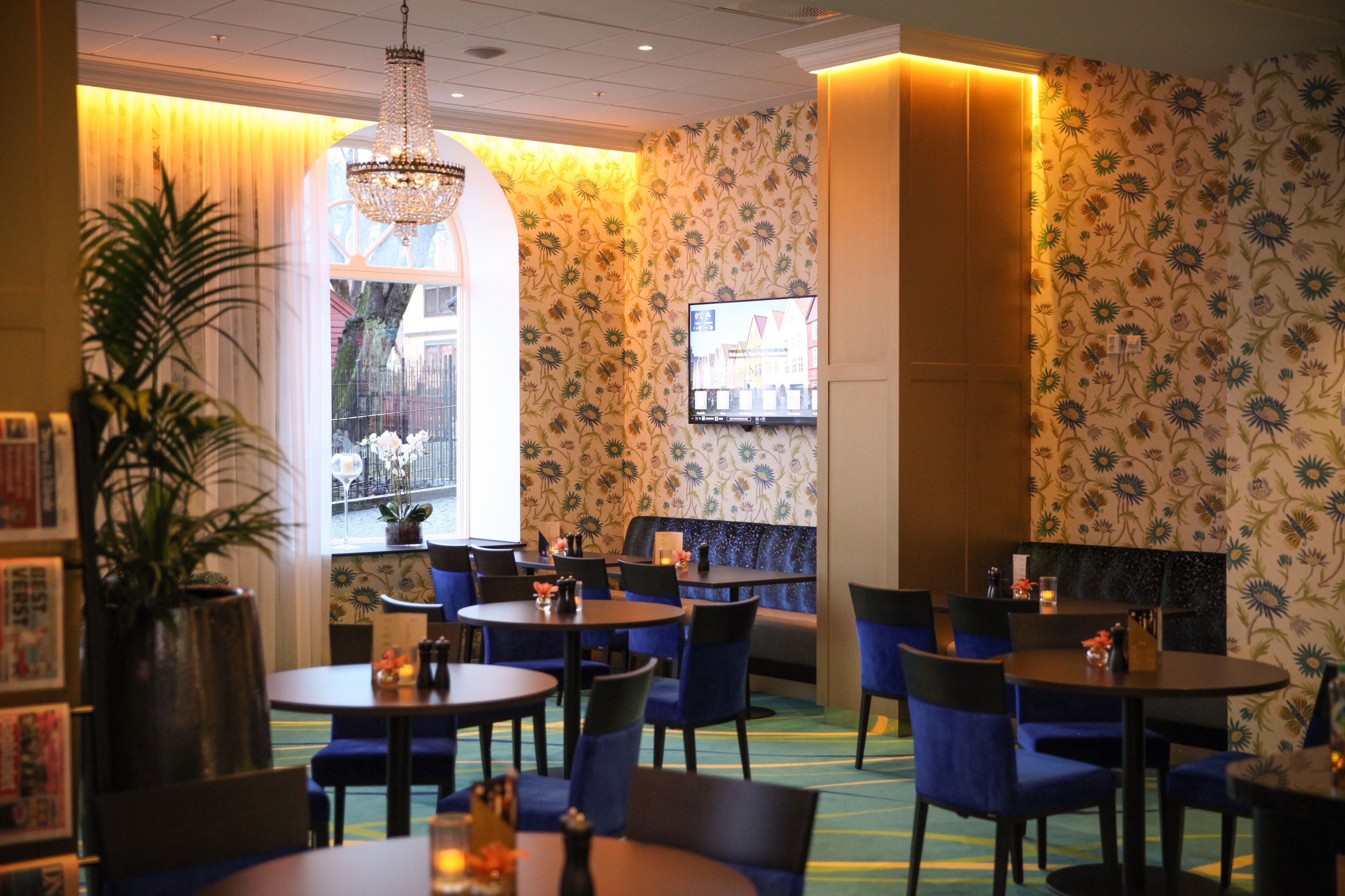 A restaurant with round tables, blue chairs, a chandelier, and floral wallpaper.