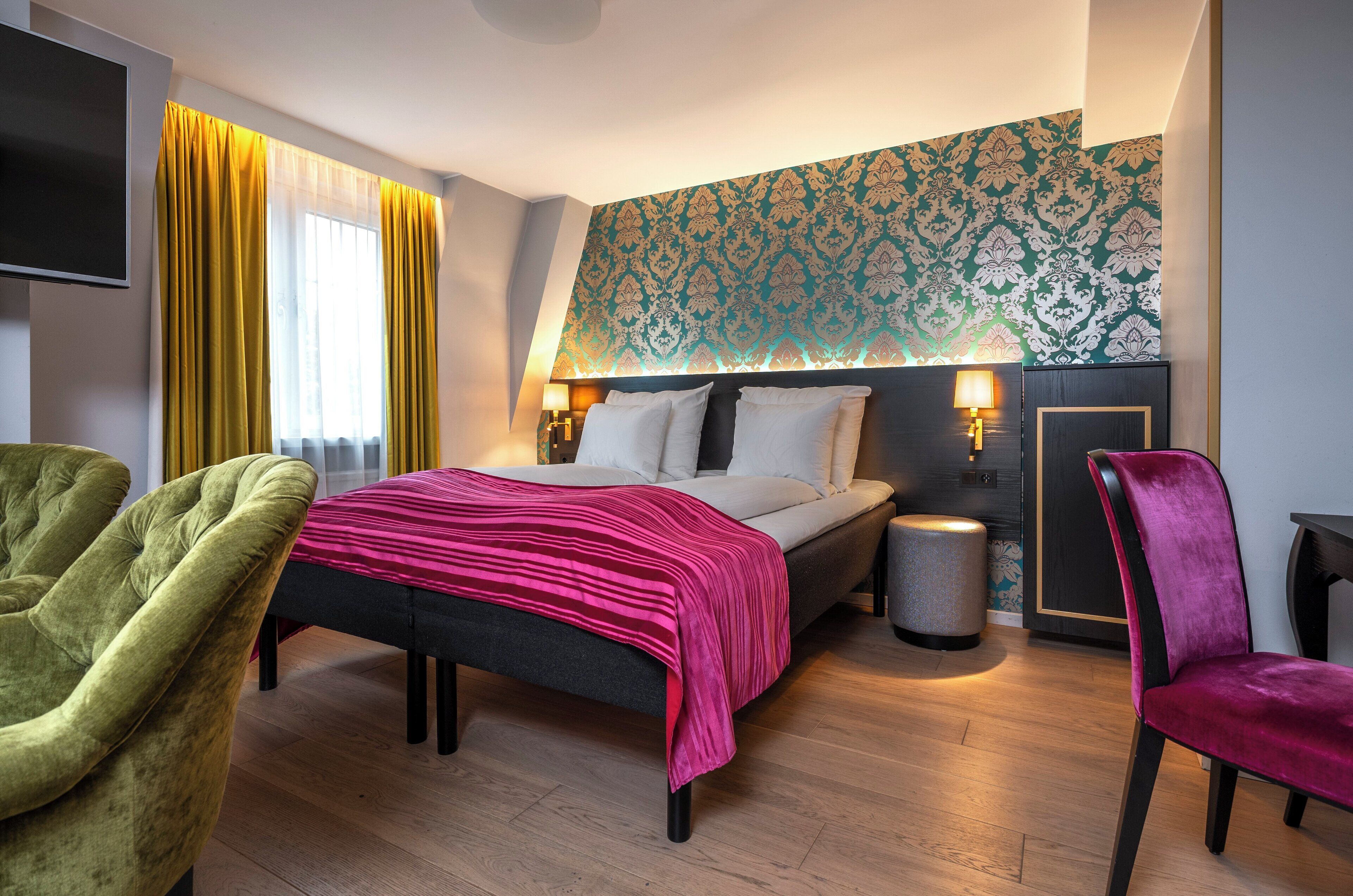 Una habitación de hotel con una cama, dos mesitas de noche con lámparas, un sillón verde, una silla rosa, una pequeña mesa redonda y un papel tapiz estampado en la pared.