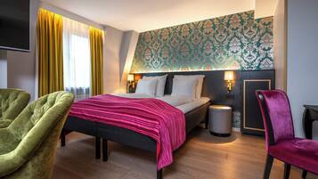 Una habitación de hotel con una cama, dos mesitas de noche con lámparas, un sillón verde, una silla rosa, una pequeña mesa redonda y un papel tapiz estampado en la pared.