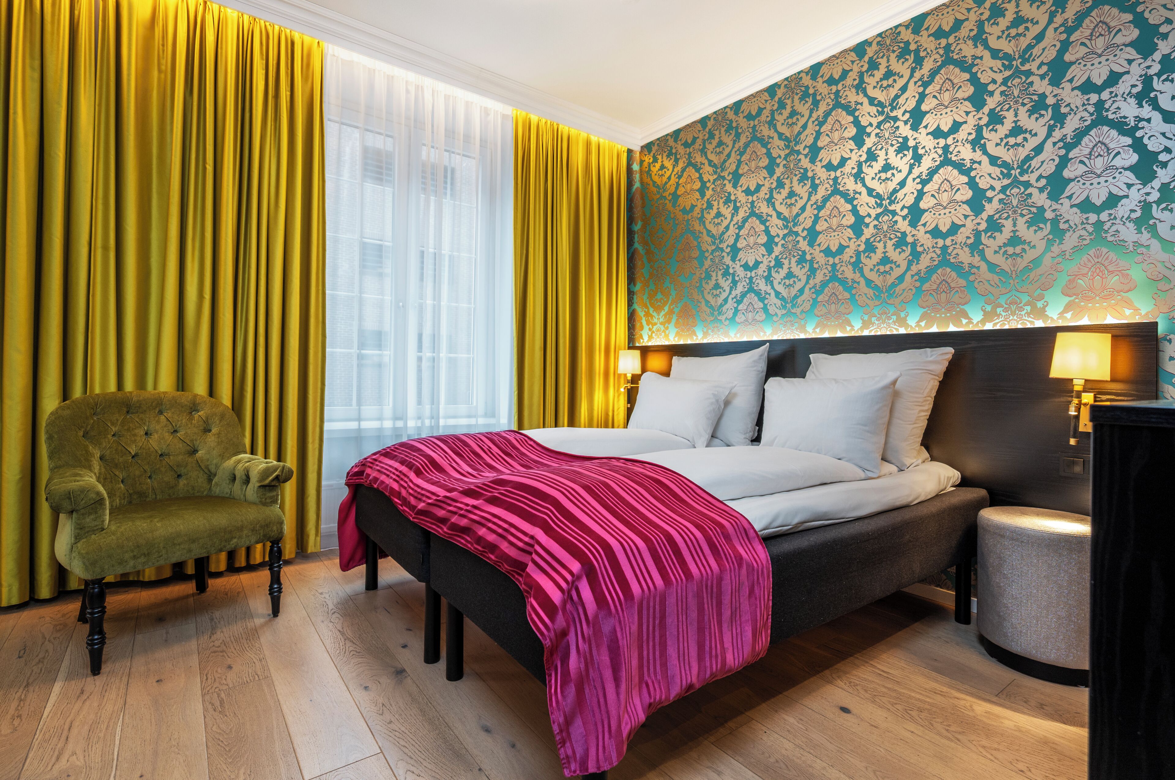A hotel room with a bed, a green armchair, a bedside table, and a patterned wall.