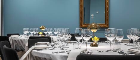 Comedor con mesas dispuestas para una comida, incluyendo copas de vino, cubiertos y un centro de mesa dorado.