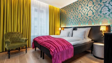 A hotel room with a bed, a green armchair, a bedside table with a lamp, and a patterned wallpaper.