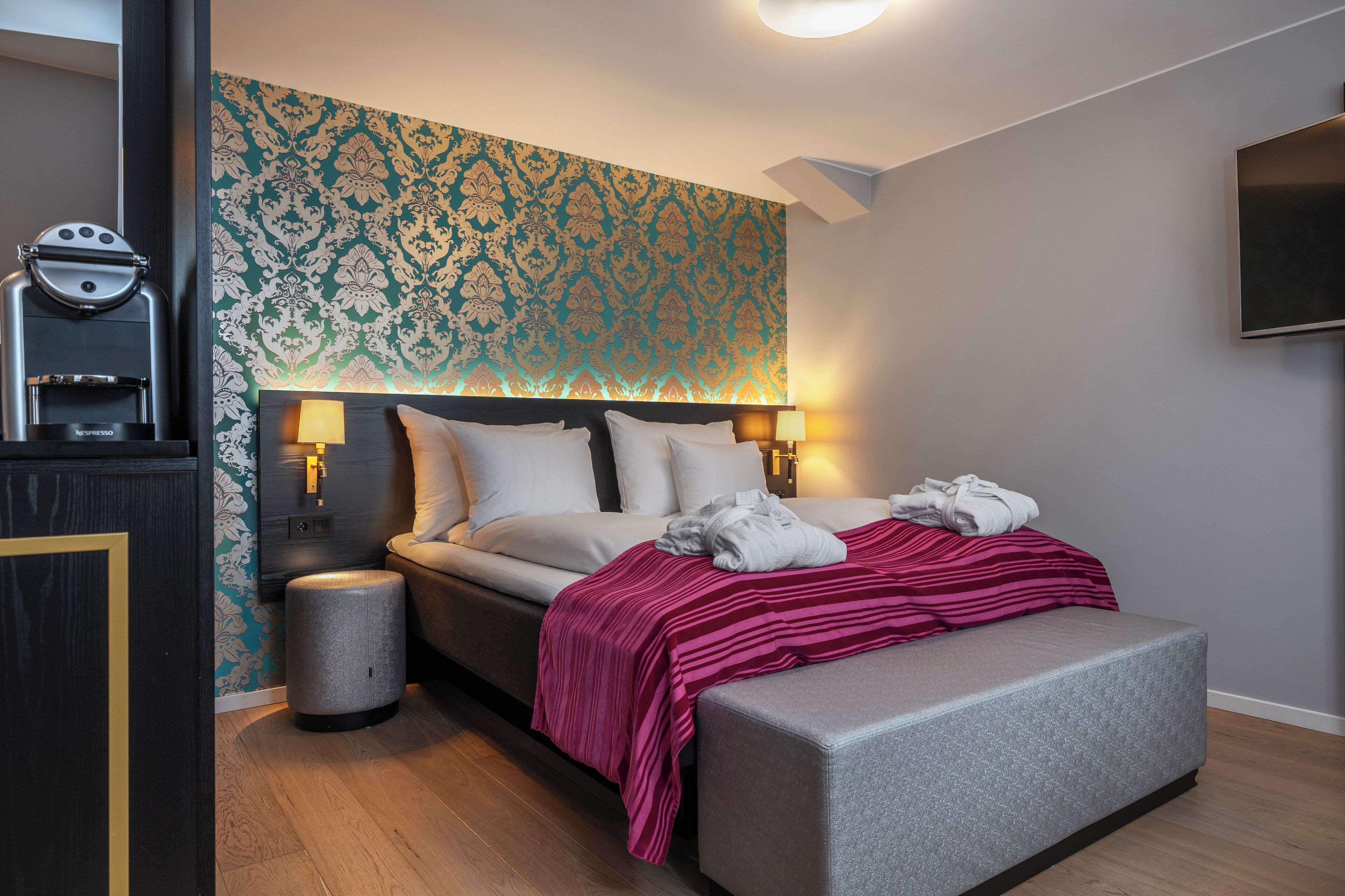 A modern hotel room with a large bed, two bedside lamps, a TV mounted on the wall, and a patterned wallpaper.