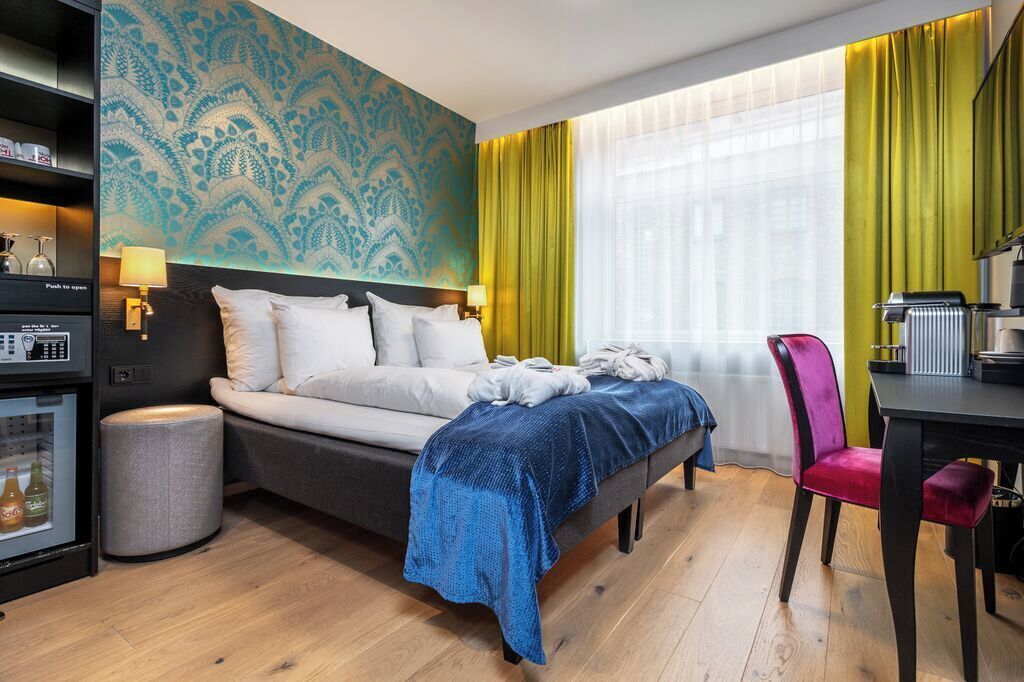 A hotel room with a bed, a desk, a chair, a minibar, and a patterned wall.