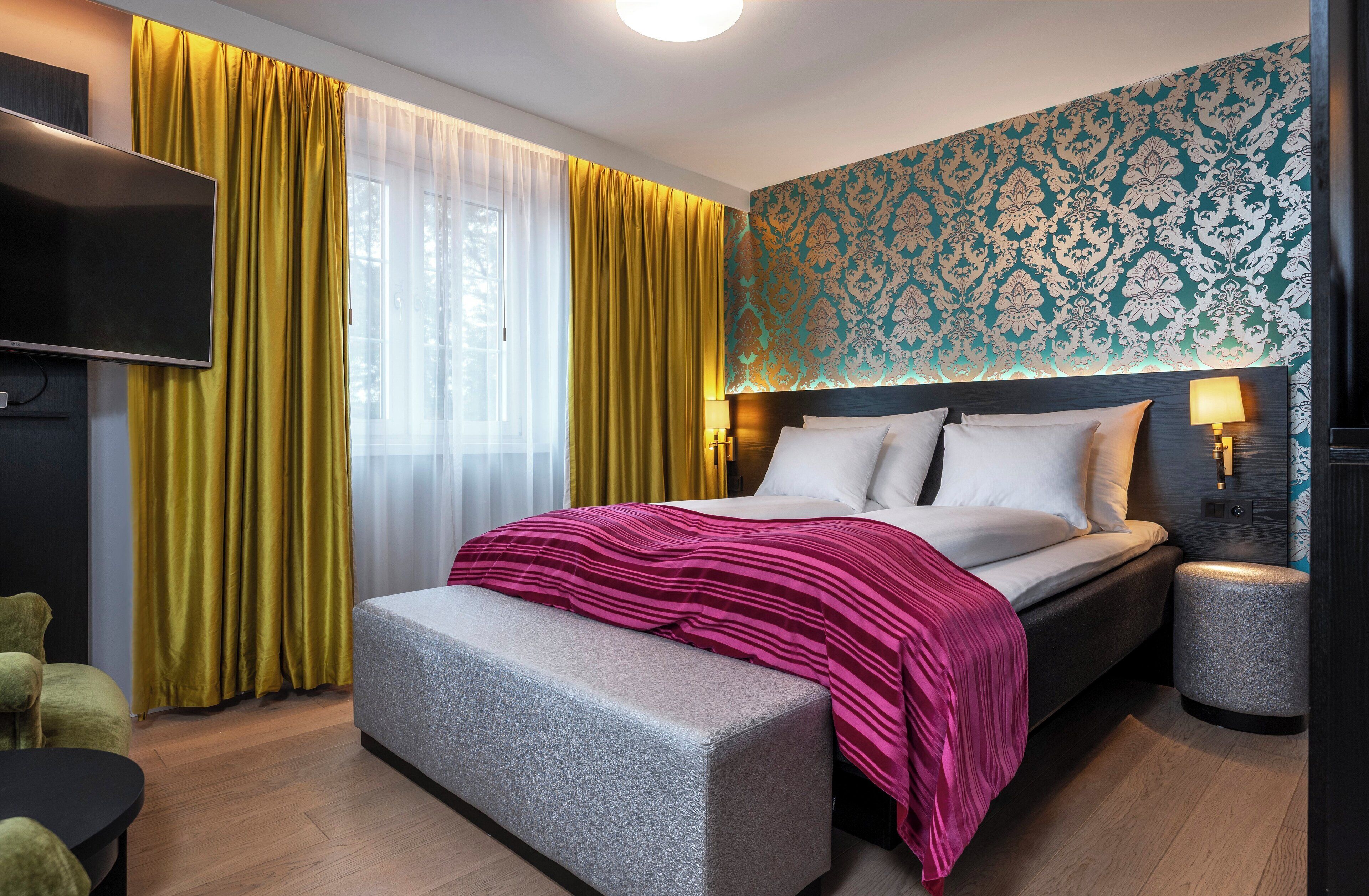 A modern hotel room with a large bed, a bench, a TV, and a patterned wall.
