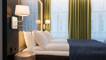 Ett modernt hotellrum med en snyggt bäddad säng, två väggmonterade lampor och ett stort fönster med gröna gardiner.