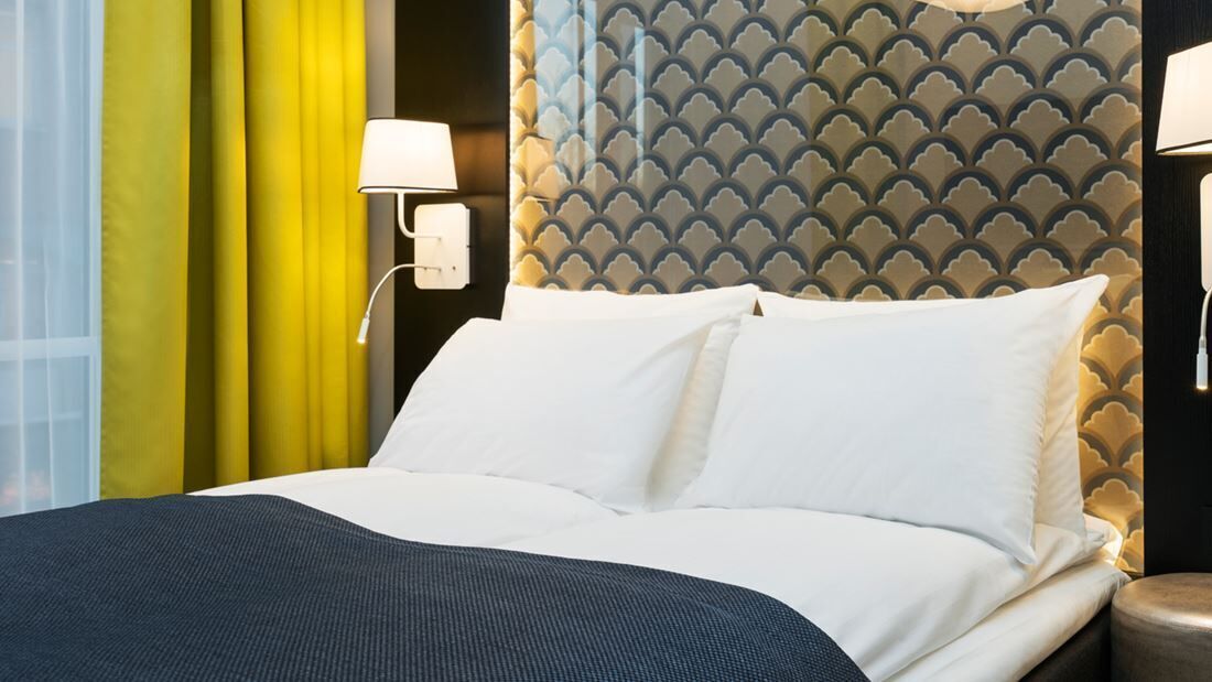 Habitación de hotel con cama, cortinas amarillas, cabecero estampado y lámparas fijadas a la pared.