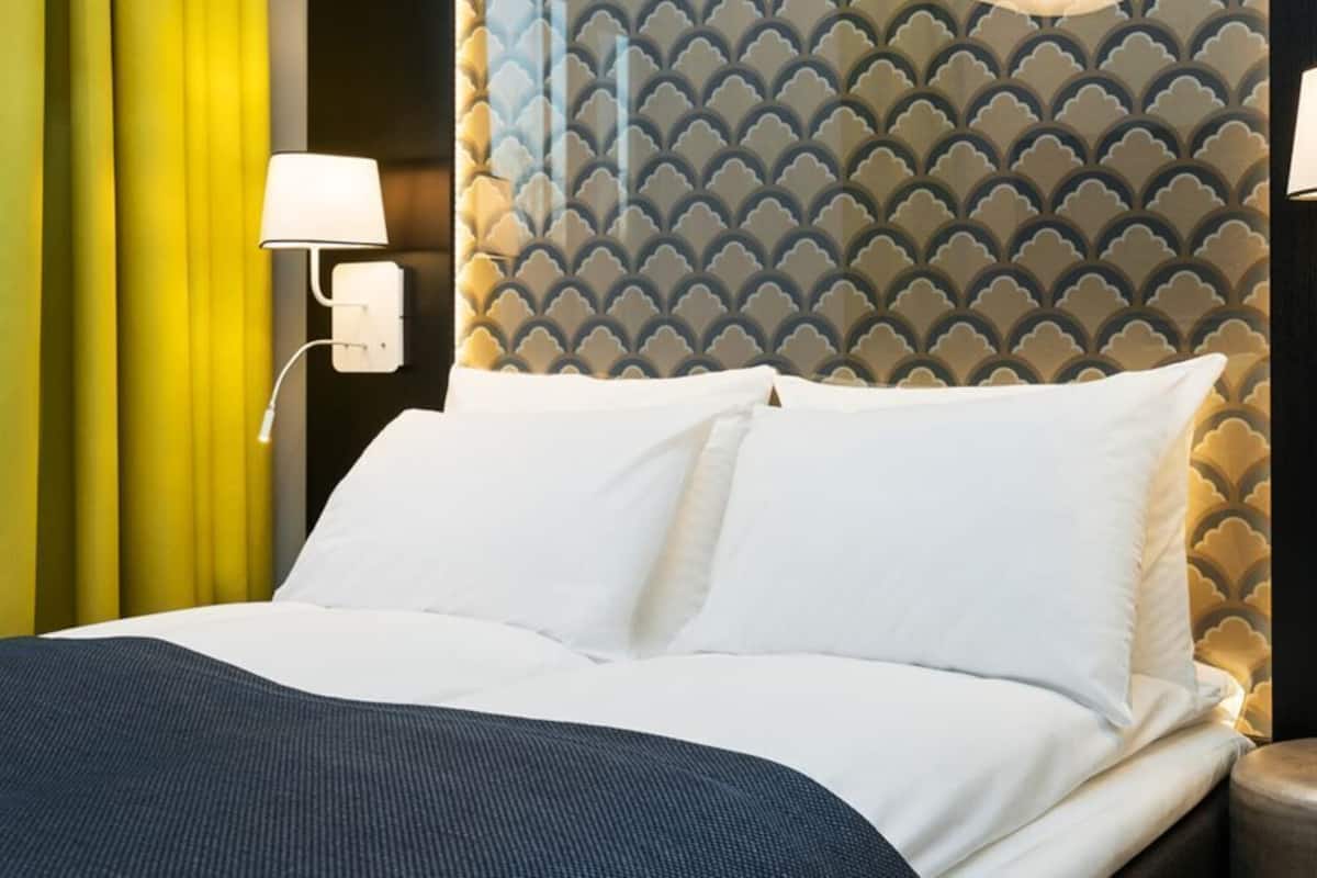 Habitación de hotel con cama, cortinas amarillas, cabecero estampado y lámparas fijadas a la pared.