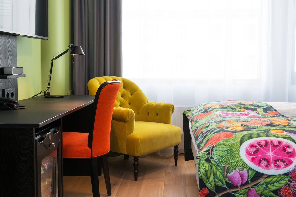 A hotel room with a desk, a yellow armchair, a bed with a colorful floral bedspread, and a window with curtains.