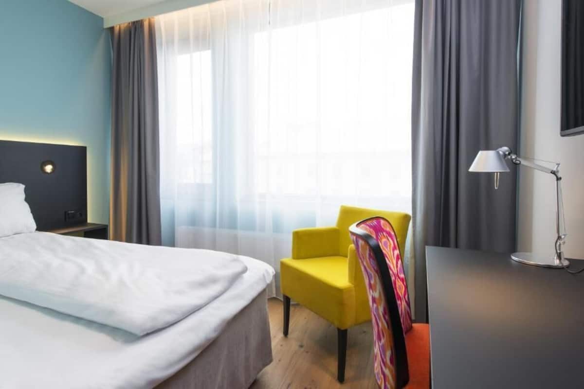 A hotel room with a bed, a chair, a desk, a lamp, and a window with curtains.