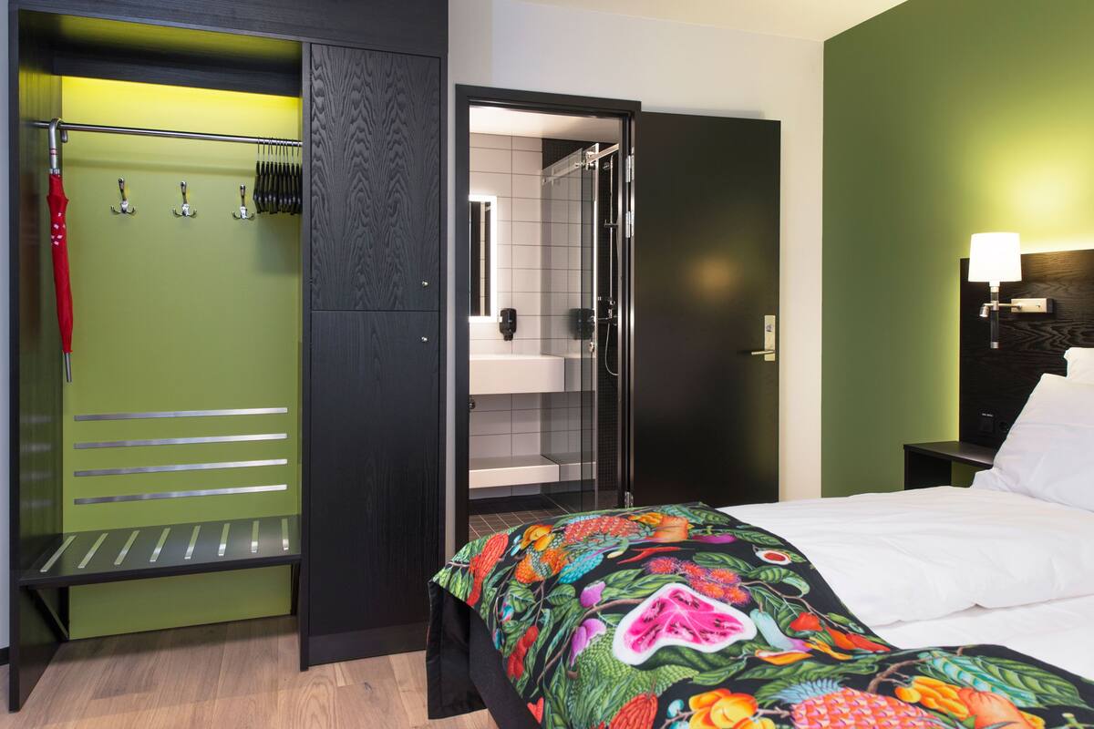 A hotel room with a green wardrobe, a shower area, a bed with a colorful blanket, and a nightstand with a lamp.