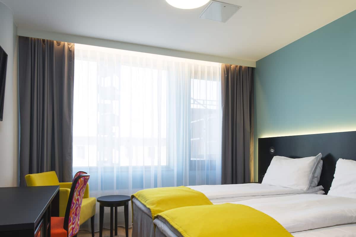 Habitación de hotel con una cama grande, un escritorio y una silla. Hay una ventana con cortinas, una lámpara en el techo y una pared con cabecera.