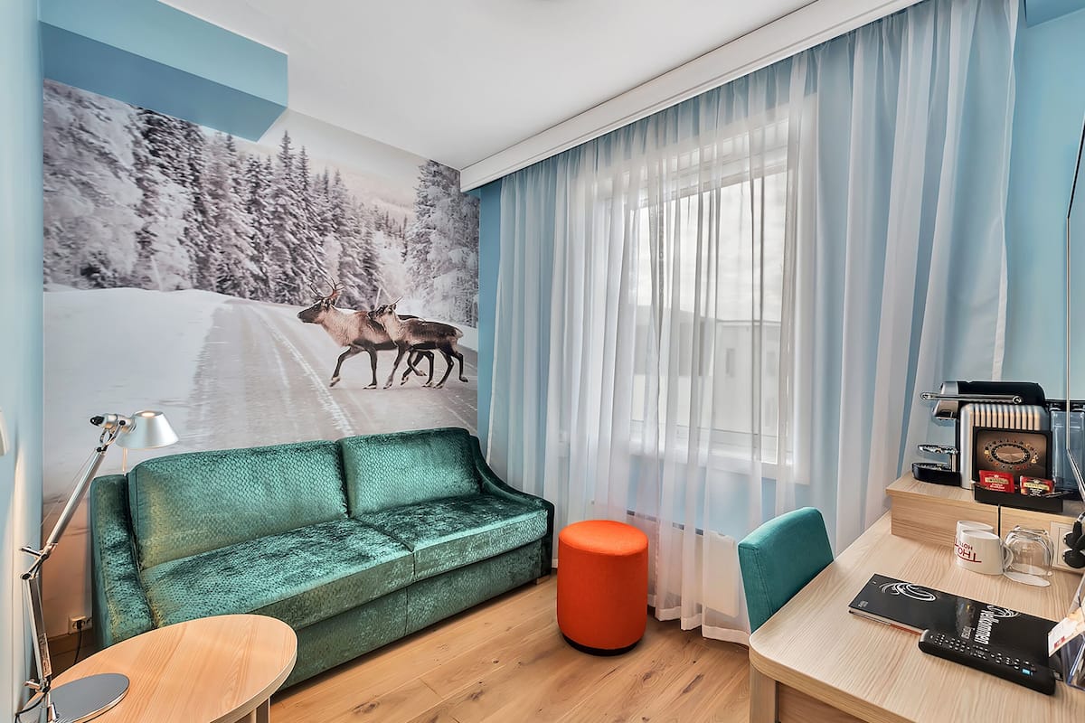 A room with a green sofa, a wooden table, a red ottoman, a desk with a computer, and a wall mural of reindeer on a snowy road.