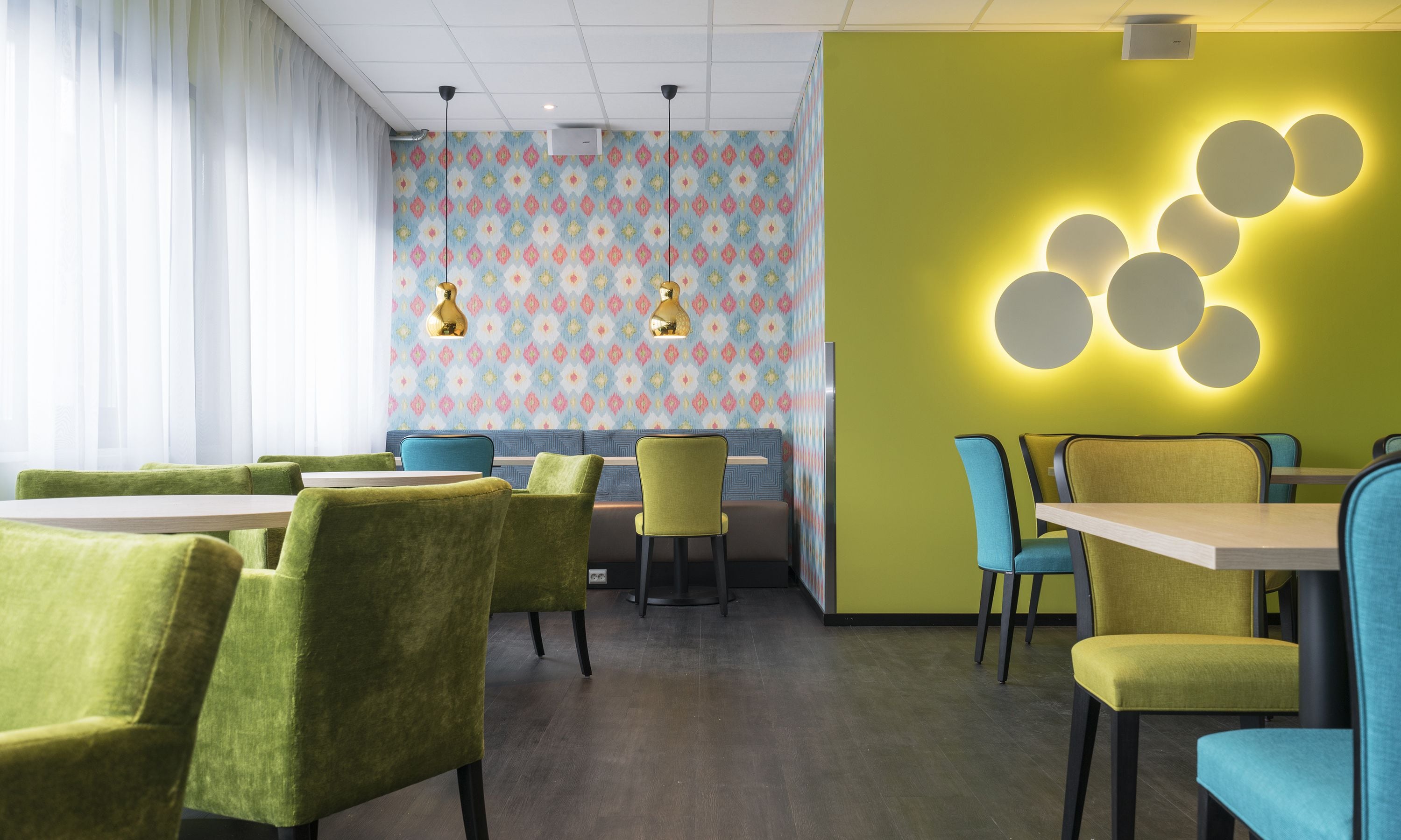 Un comedor moderno con sillas tapizadas en verde y azul, una pared con diseño y una lámpara circular original.