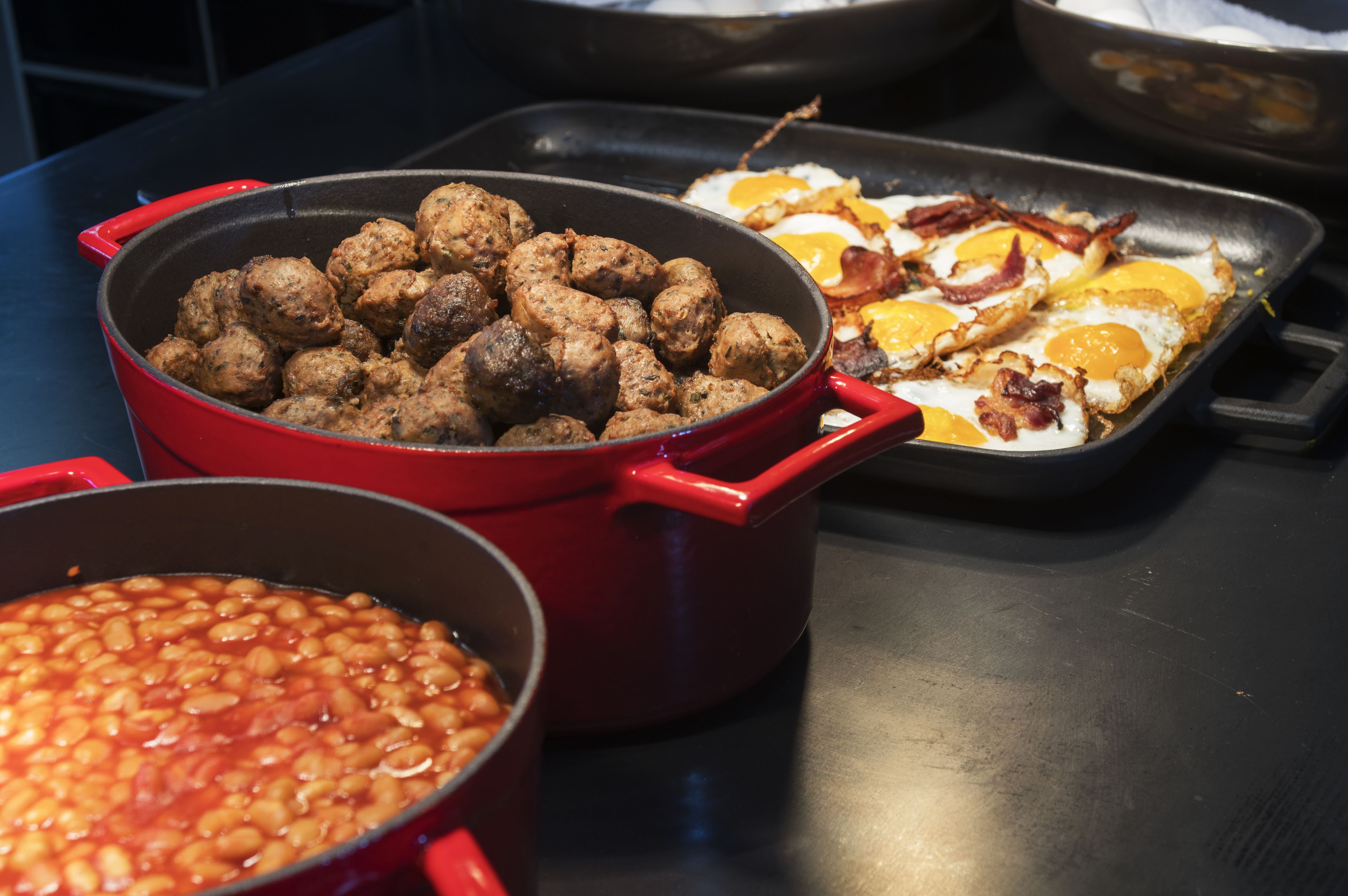 A kitchen counter with three cooking pots: one with meatballs, one with baked beans, and one with fried eggs and bacon.