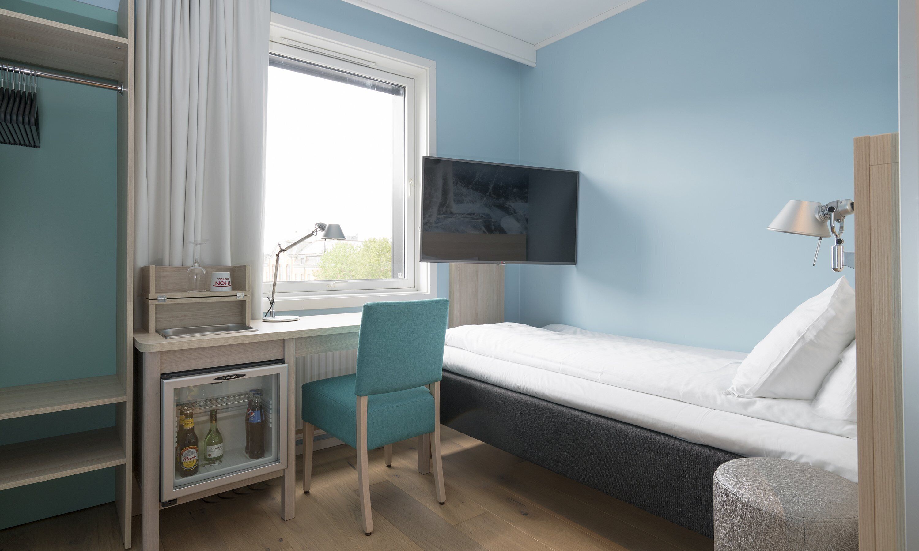 A compact room with a bed, a desk with a chair, a television mounted on the wall, and a small refrigerator.