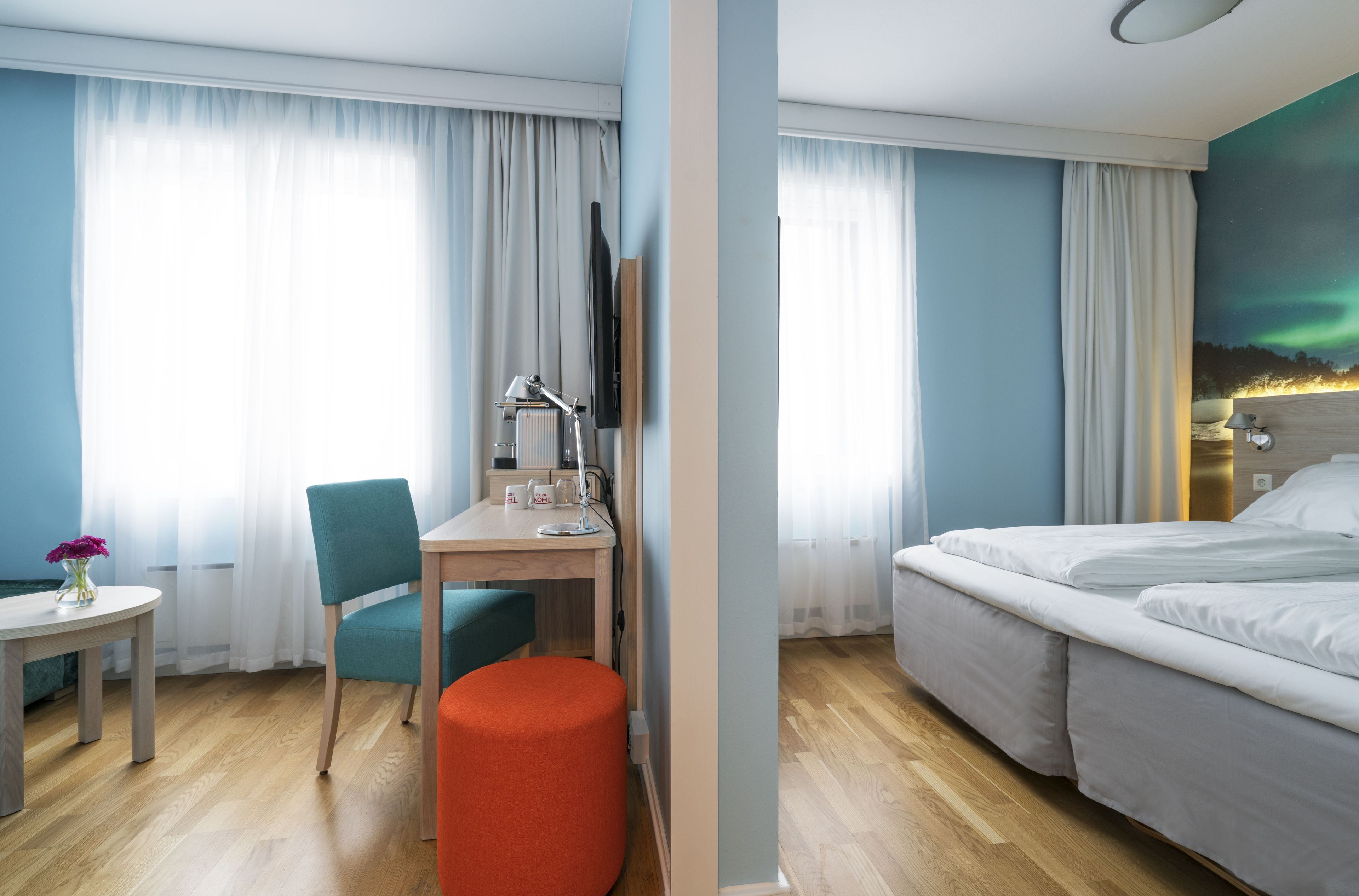 Habitación de hotel con una cama, un escritorio con una silla, una mesita con un jarrón de flores y una ventana con cortinas.