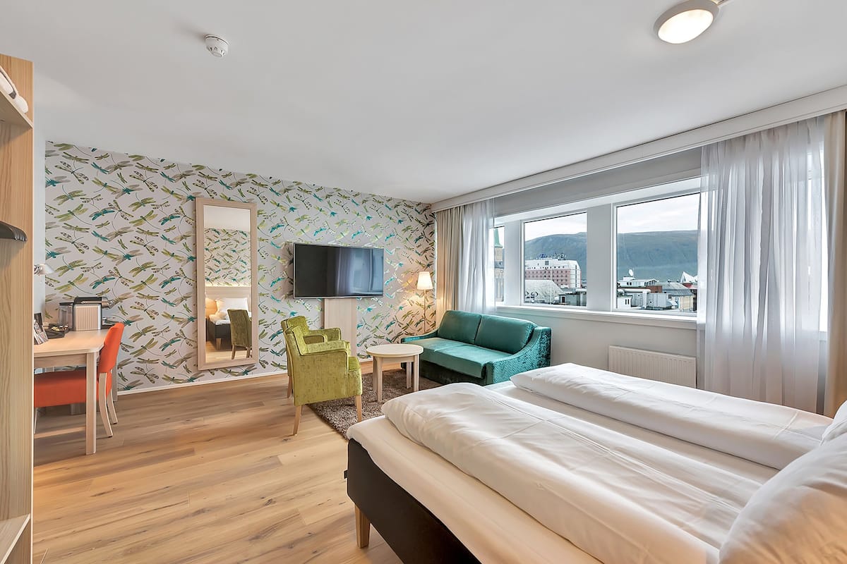 A hotel room with a bed, a desk with a chair, a television, and a view of the city through large windows.