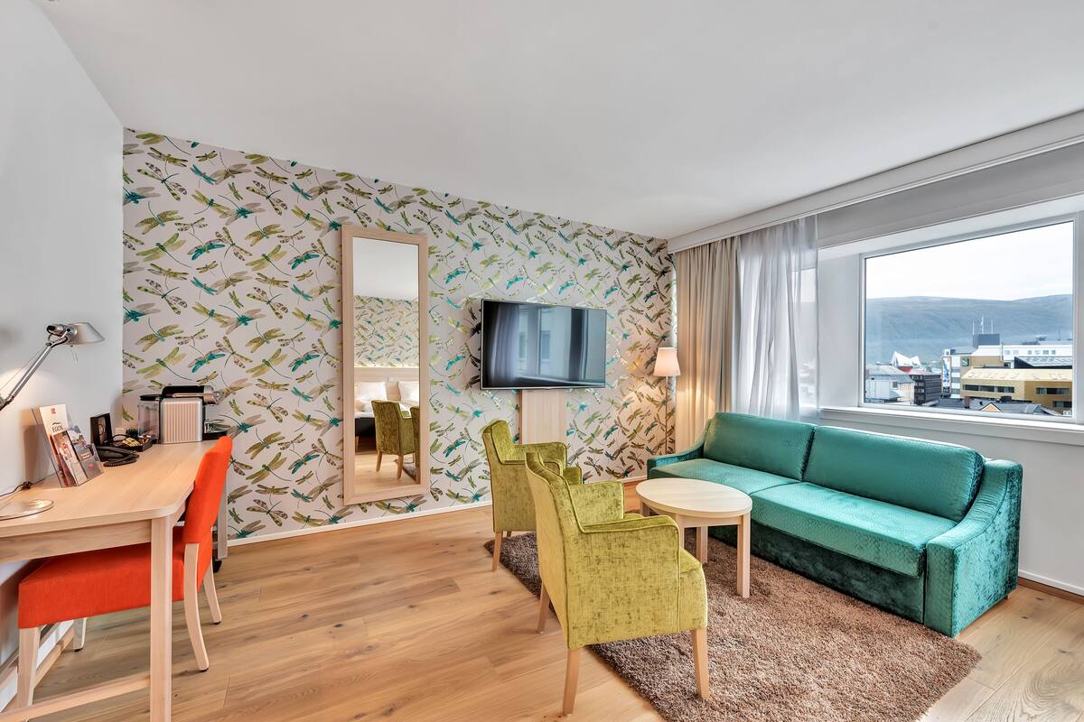 En moderne stue med en grøn sofa, et spisebord med stole, et fjernsyn og et stort vindue med udsigt til bygninger og bjerge.