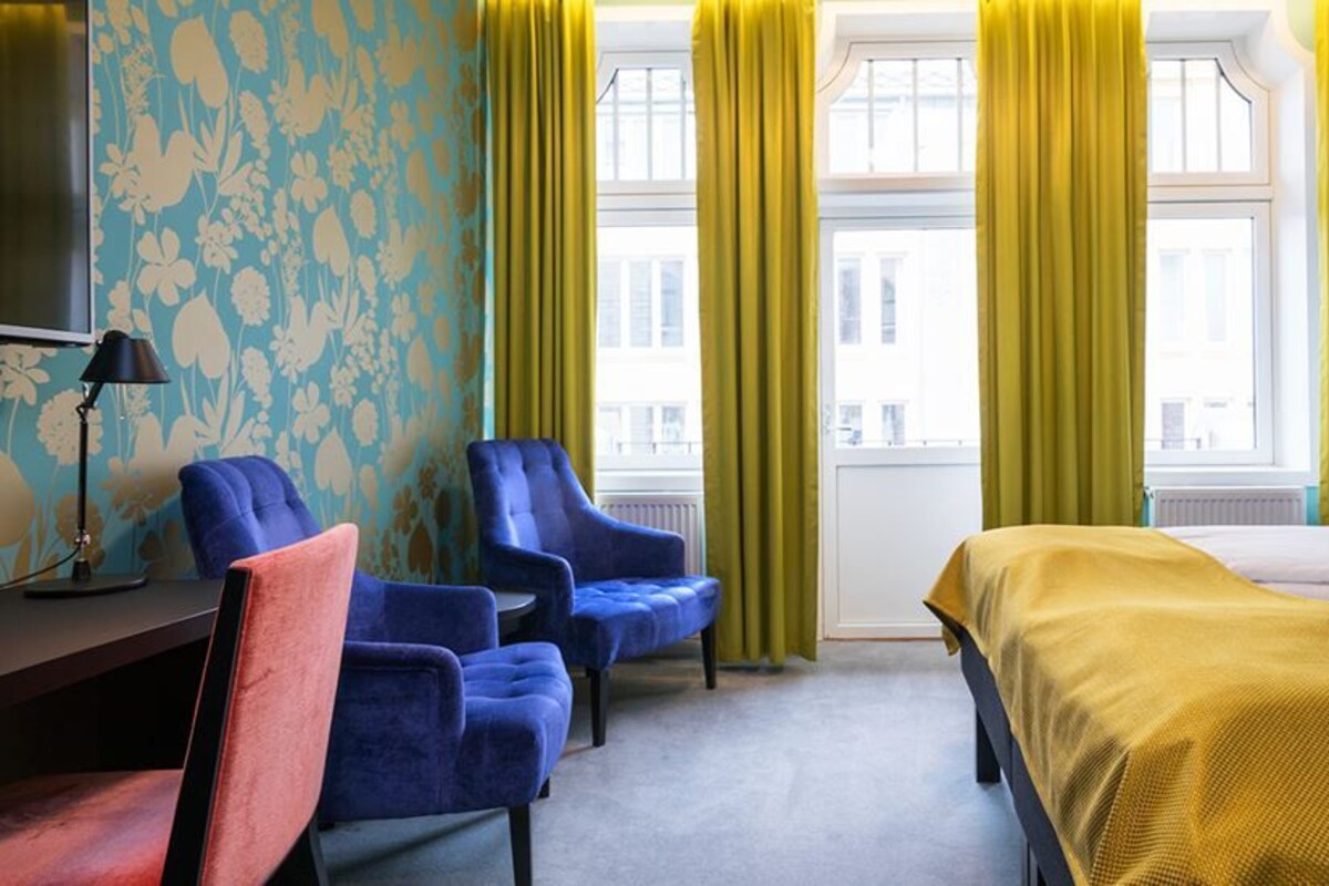 Habitación de hotel con una cama, dos sillones azules, una silla roja, un escritorio con lámpara y un televisor.