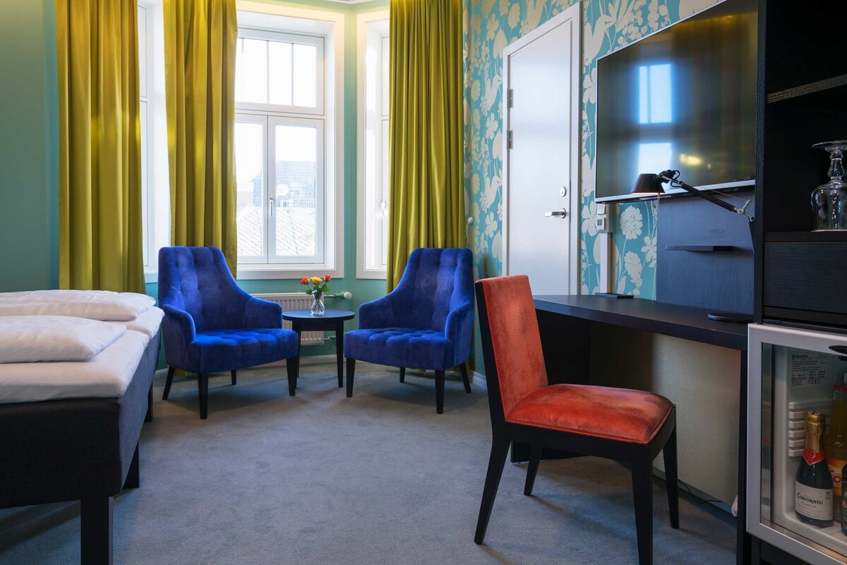 A hotel room with two beds, a desk with a television, two blue chairs, a red chair, and a minibar.