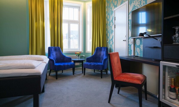 Habitación de hotel con dos camas, un escritorio con televisor, dos sillas azules y una silla roja, además de un minibar.