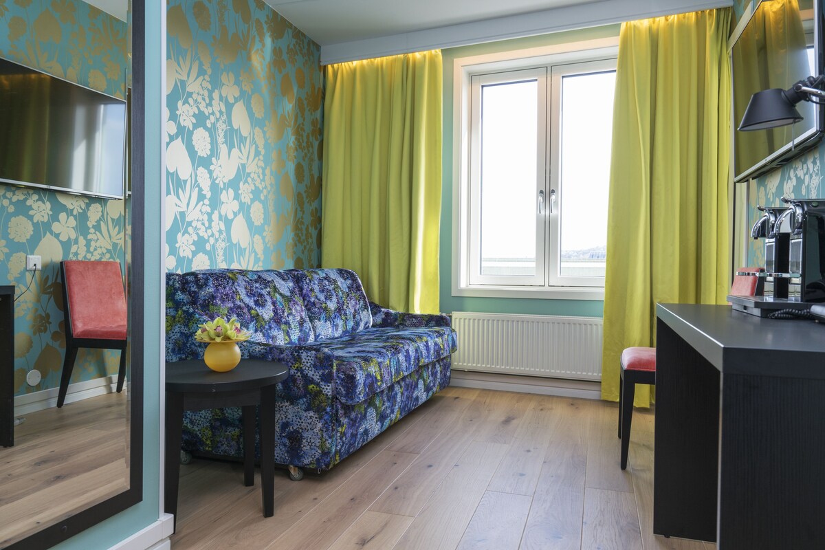 A room with a sofa, a small table with a vase, a red chair, a desk, and a window with curtains.
