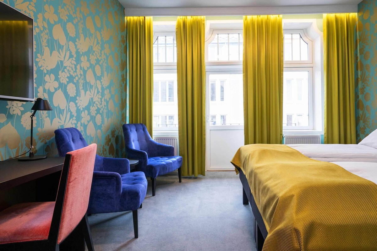 A hotel room with a bed, two armchairs, a desk with a lamp, and a TV.