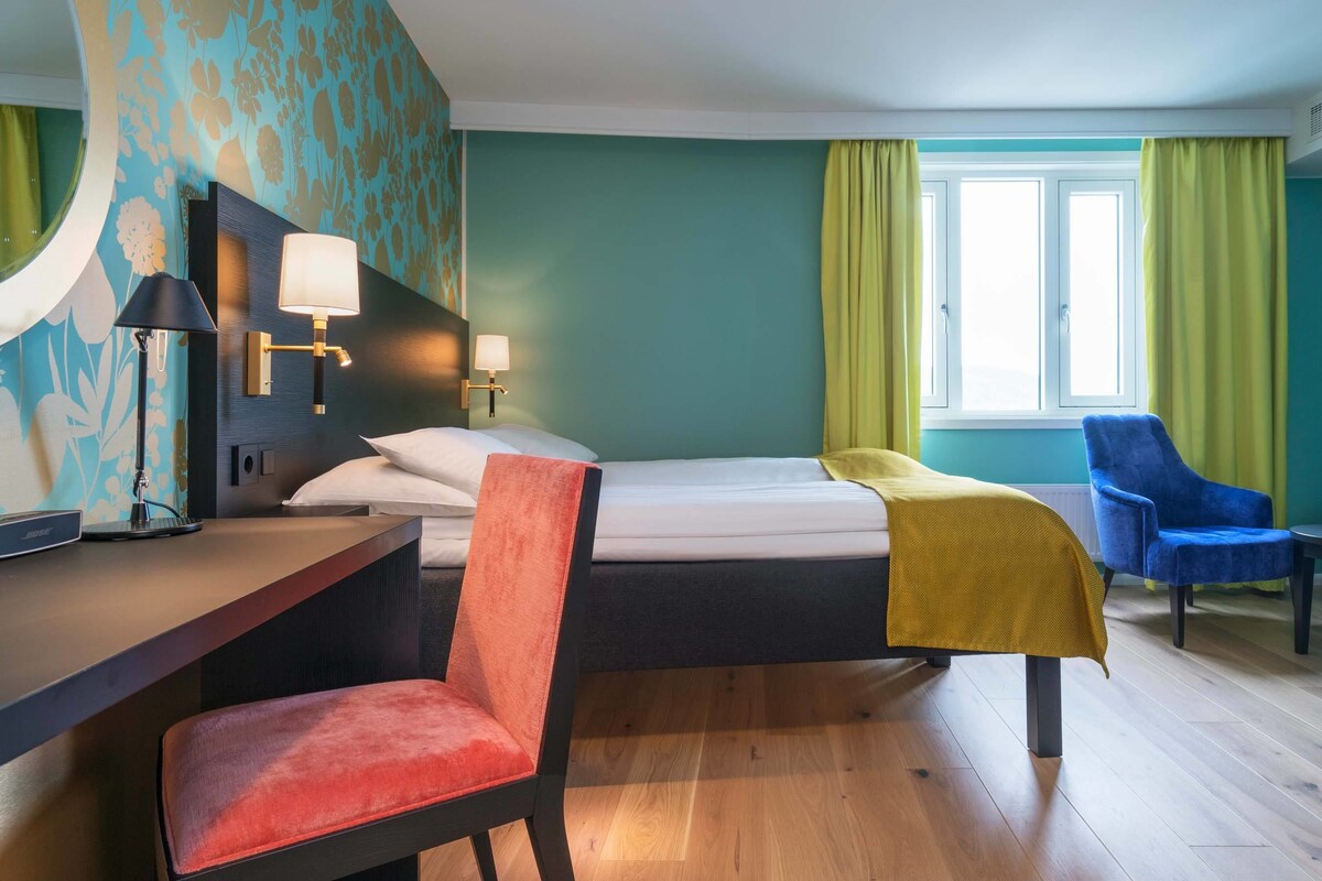 A hotel room with a bed, a desk with a lamp, a chair, a window with curtains, and a blue armchair.