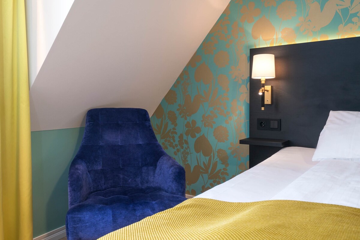 A hotel room with a bed, a blue armchair, a nightstand with a lamp, and a wall with floral wallpaper.