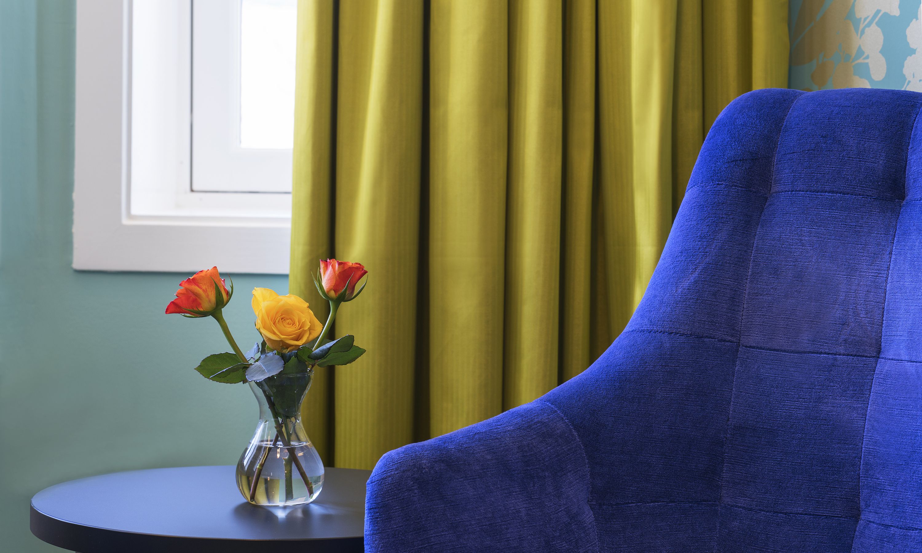 Ein Zimmer mit blauem Sessel, einem kleinen Tisch mit Blumenvase und einem Fenster mit Vorhängen.