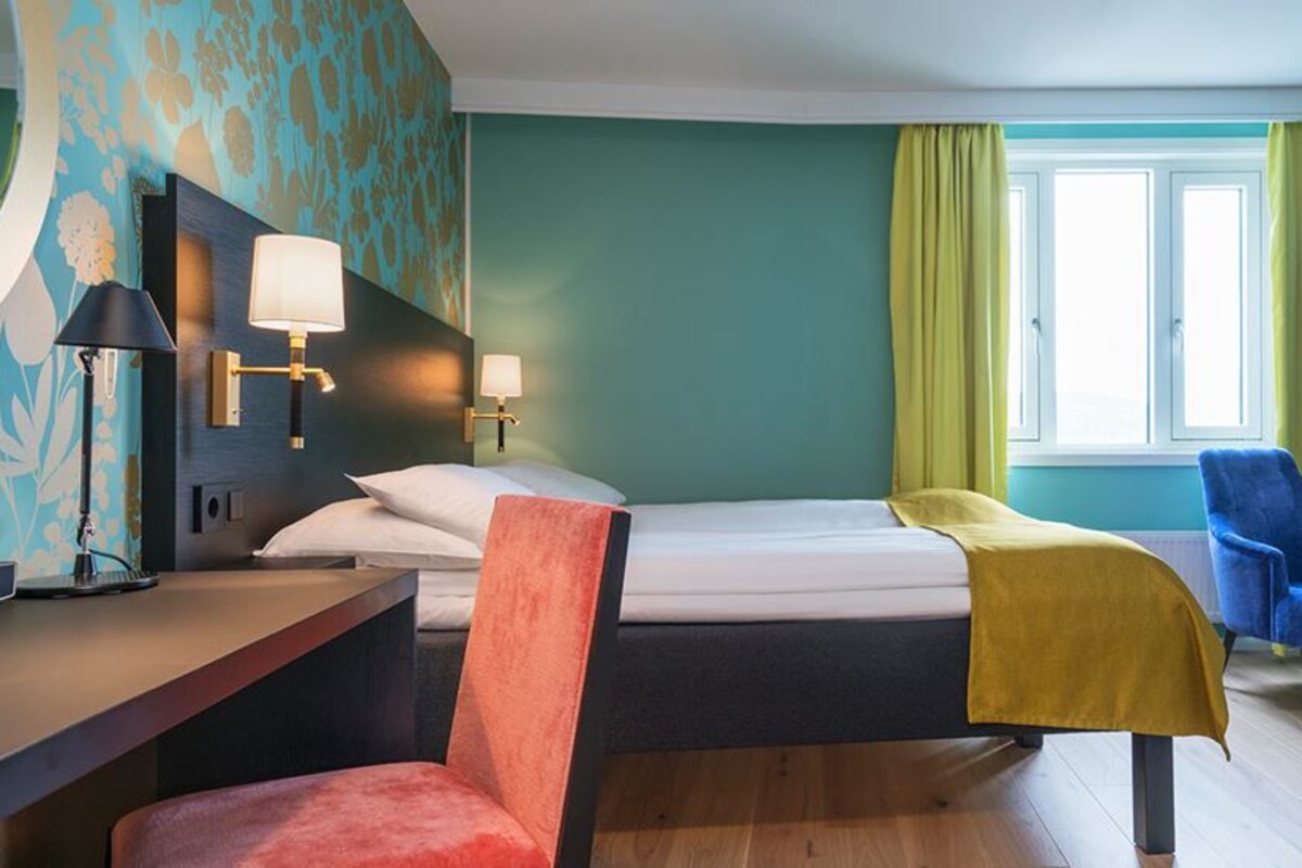Una habitación de hotel con cama, escritorio, silla y ventana con cortinas.