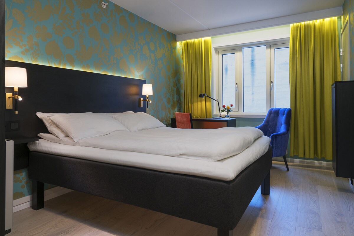 A hotel room with a large bed, a desk with a chair, a window with curtains, and a wall-mounted TV.
