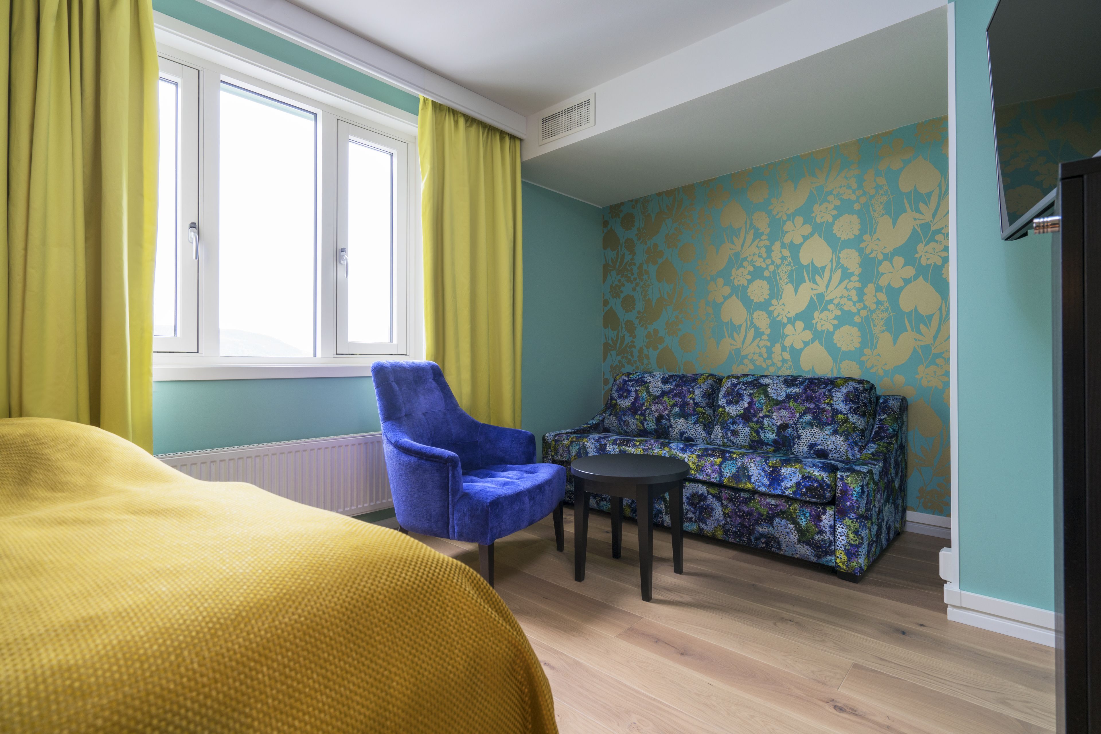 Quarto com cama, poltrona azul, sofá estampado, mesa redonda pequena e janela com cortinas amarelas.