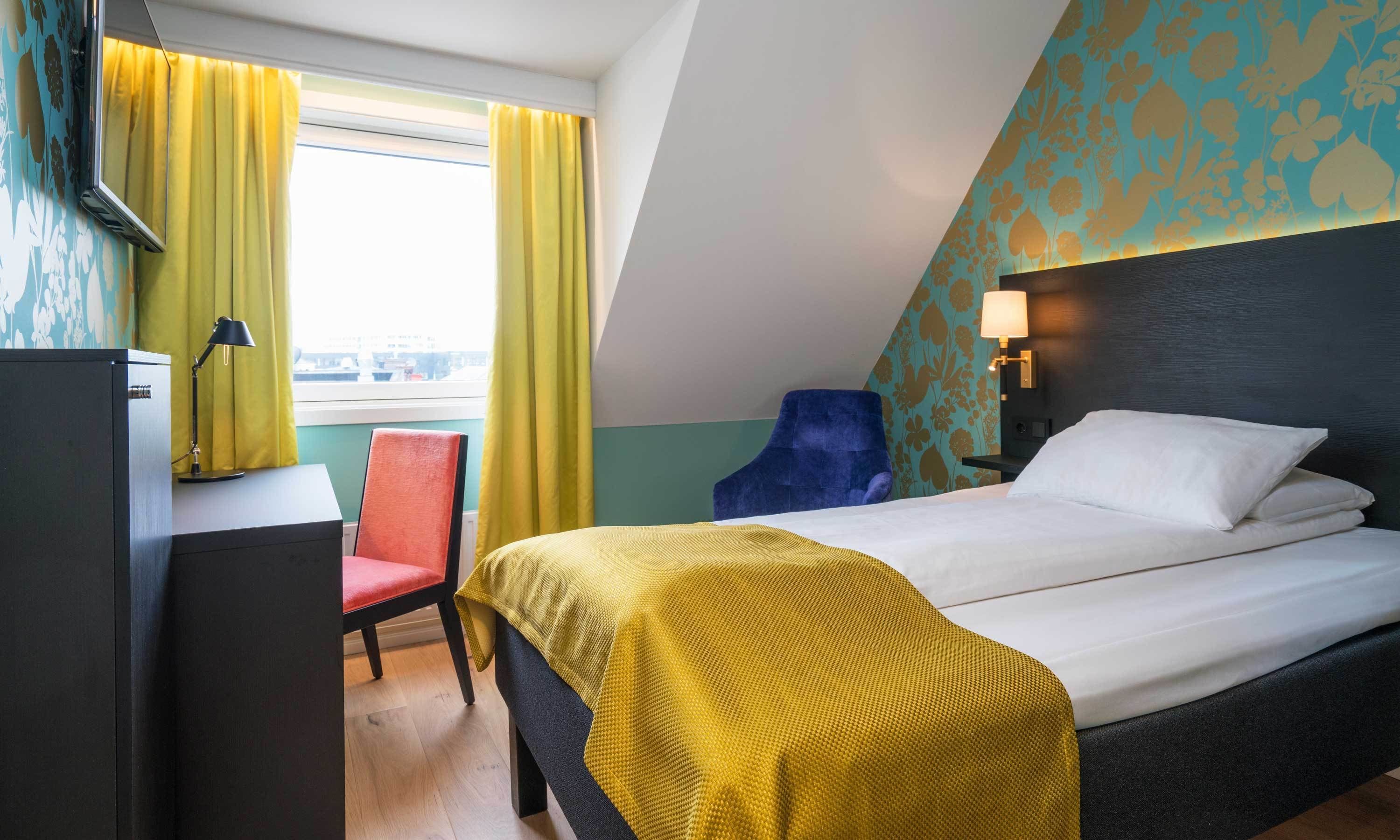 Habitación de hotel con cama, escritorio, silla, lámpara y ventana con cortinas.