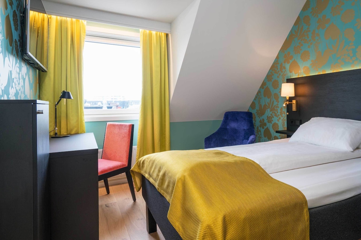 A hotel room with a bed, a desk, a chair, a lamp, and a window with curtains.