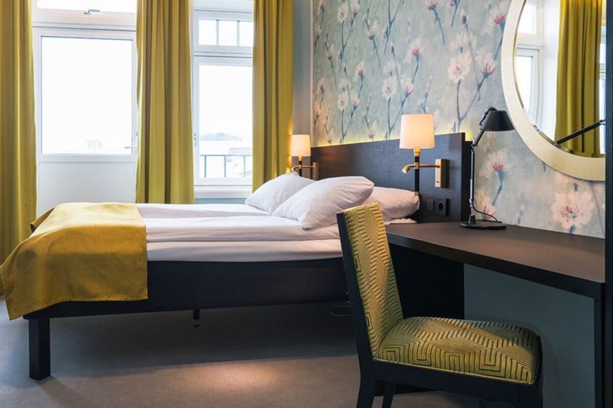 Una habitación de hotel con cama, escritorio, silla, lámpara y espejo.