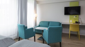 Una habitación de hotel moderna con sillas color turquesa, un sofá, un escritorio con un televisor de pantalla plana y una lámpara.