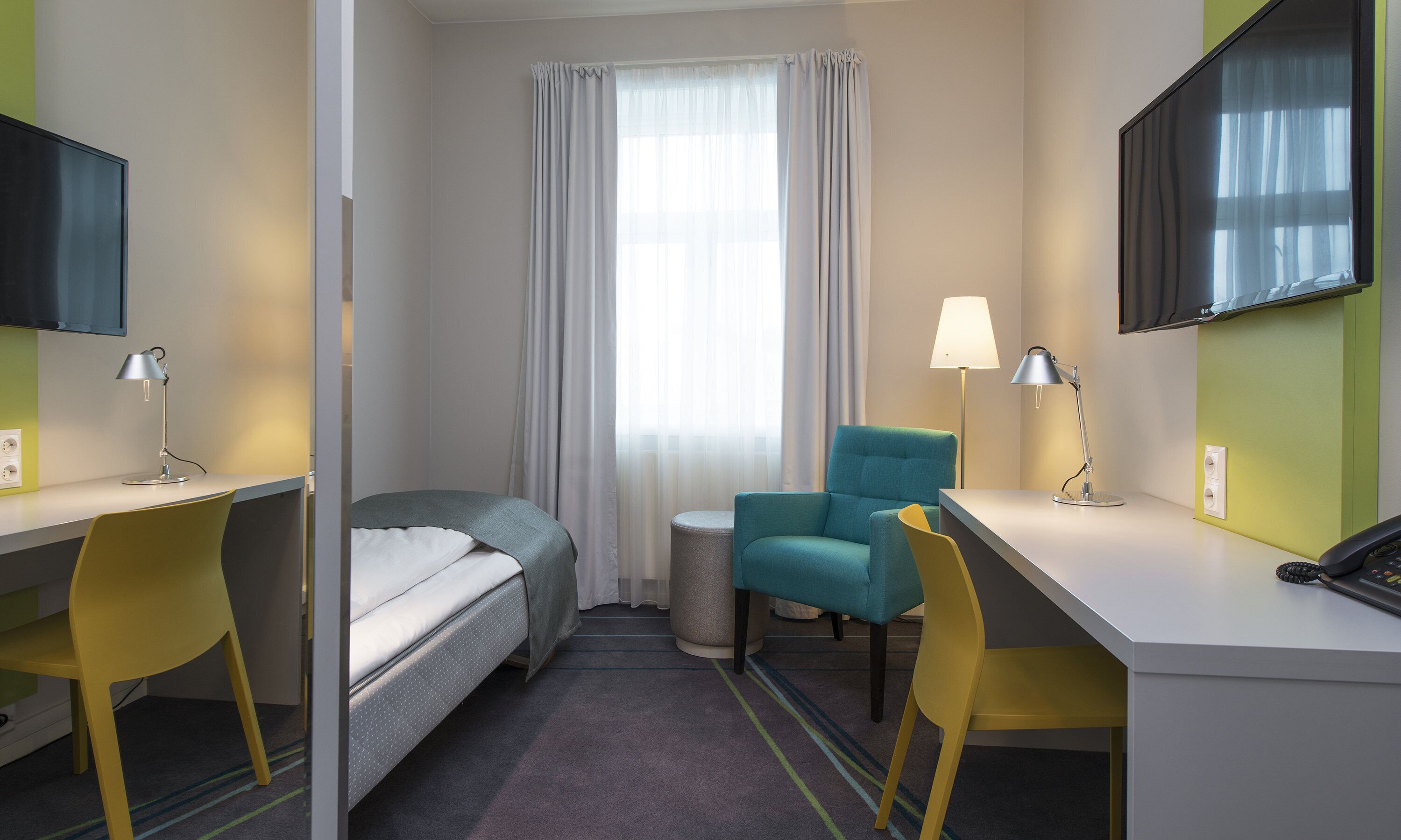 Una habitación de hotel con cama, dos sillas, escritorio, lámpara, televisión y ventana con cortinas.