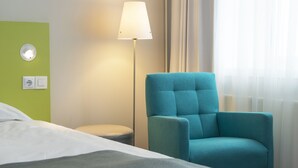 Una habitación de hotel con una cama, un sillón verde azulado, una lámpara de pie, un enchufe de pared y una toma de corriente.