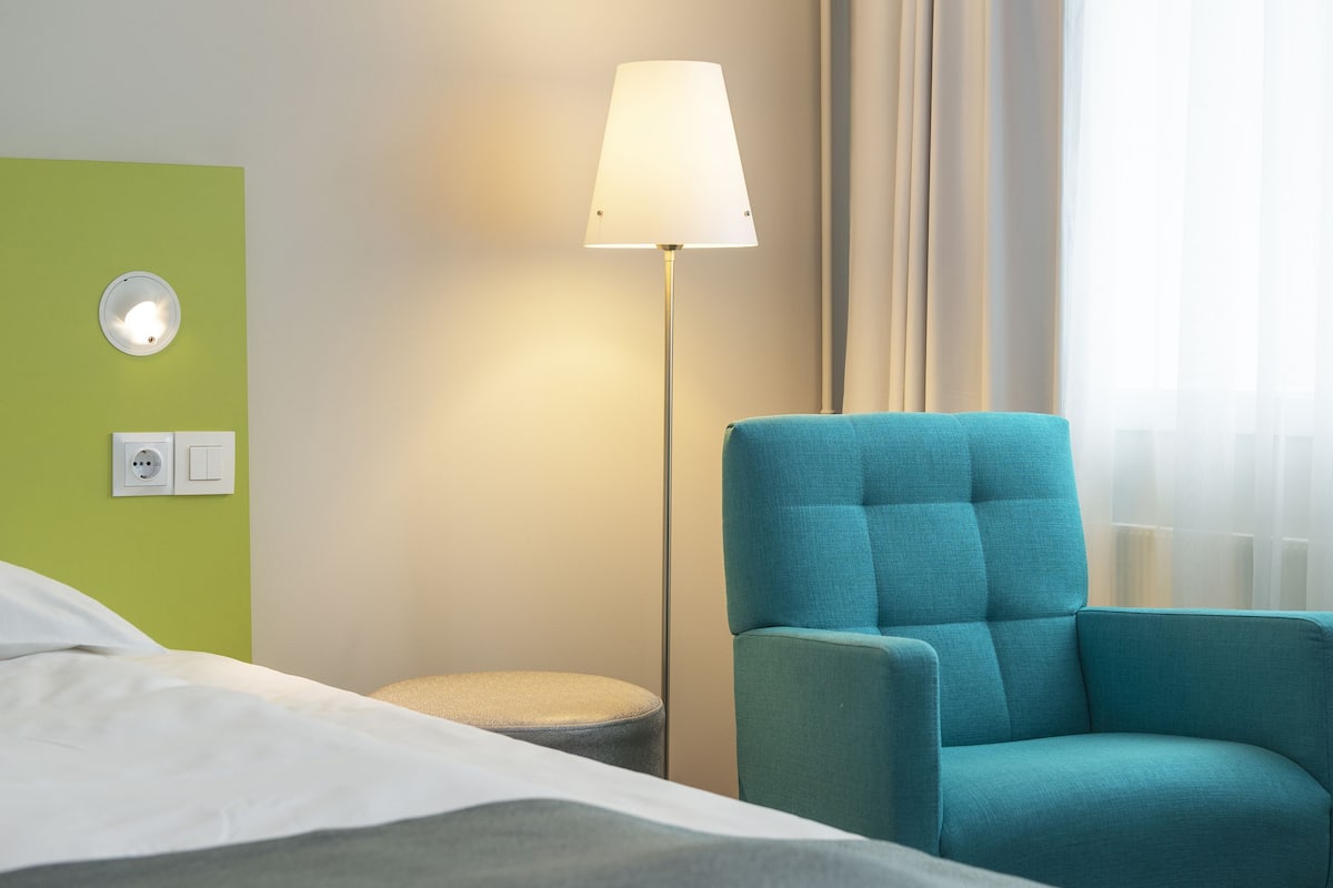 A hotel room with a bed, a teal armchair, a floor lamp, a wall socket, and a power outlet.
