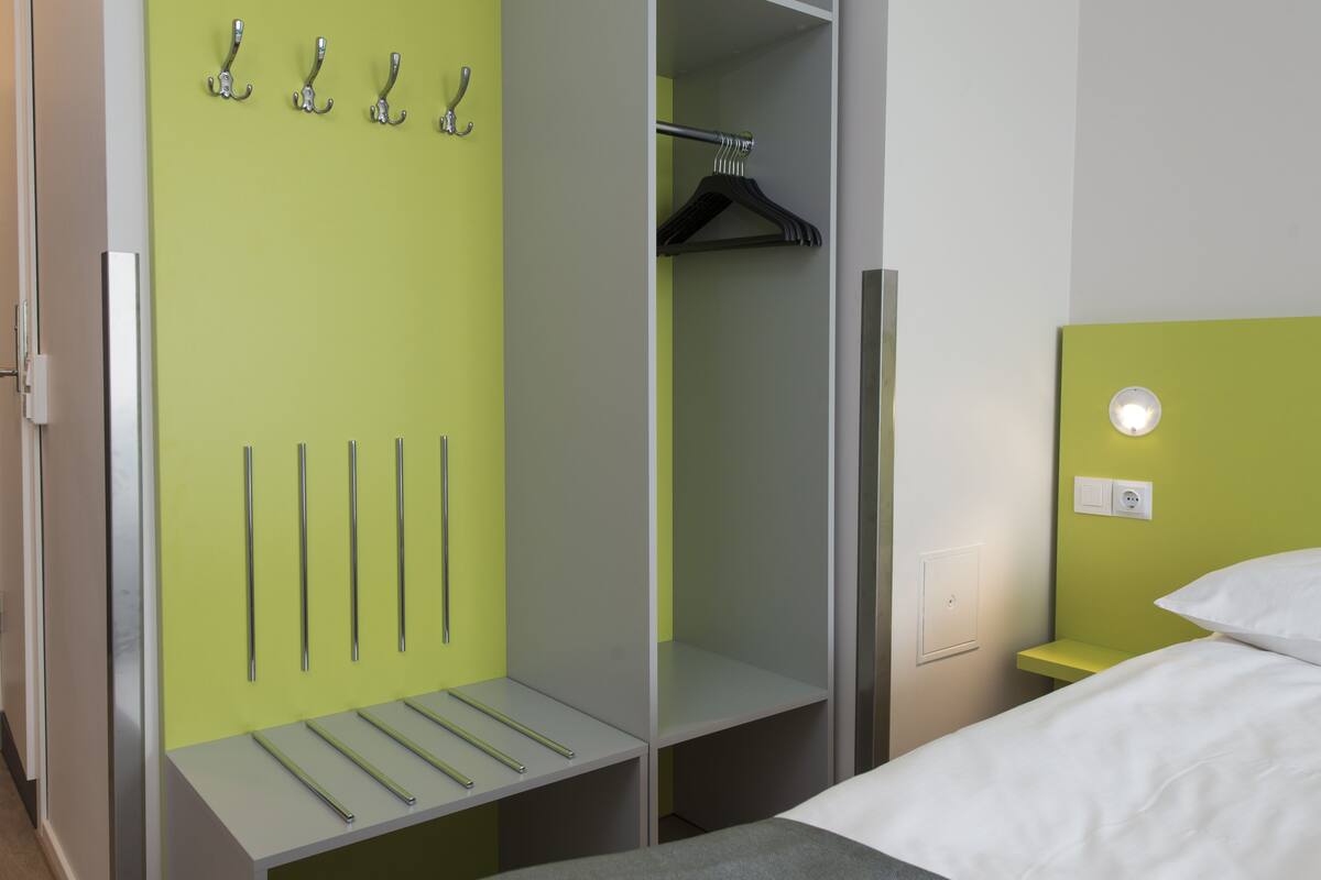 A hotel room with a bed, a wardrobe with hooks and a hanging rack, and a bedside table with a light.