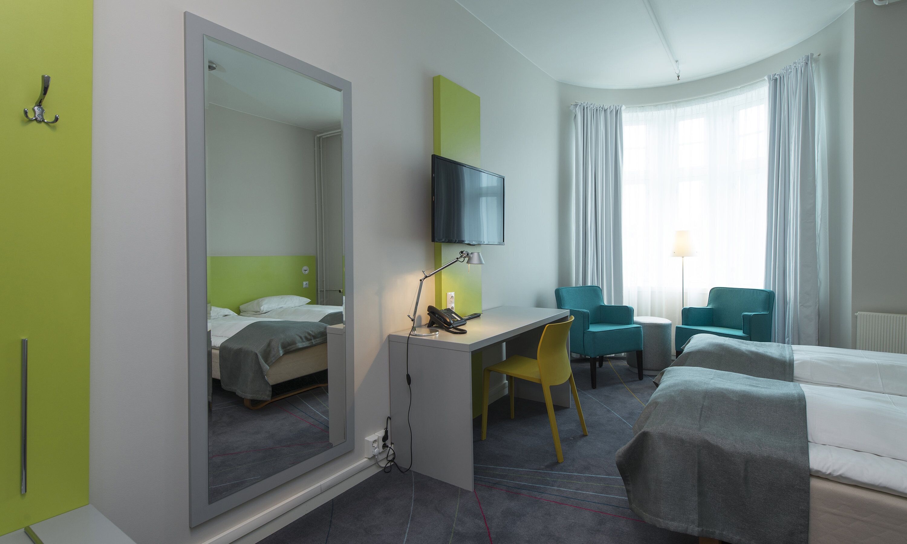 Quarto de hotel com cama, escrivaninha com cadeira, televisão e janela com cortinas.