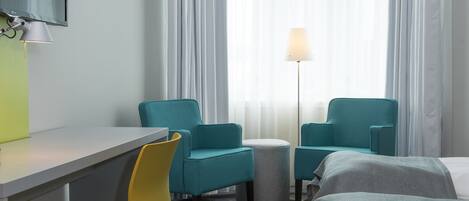 A hotel room with a desk, two teal chairs, a small round table, a lamp, and two beds.