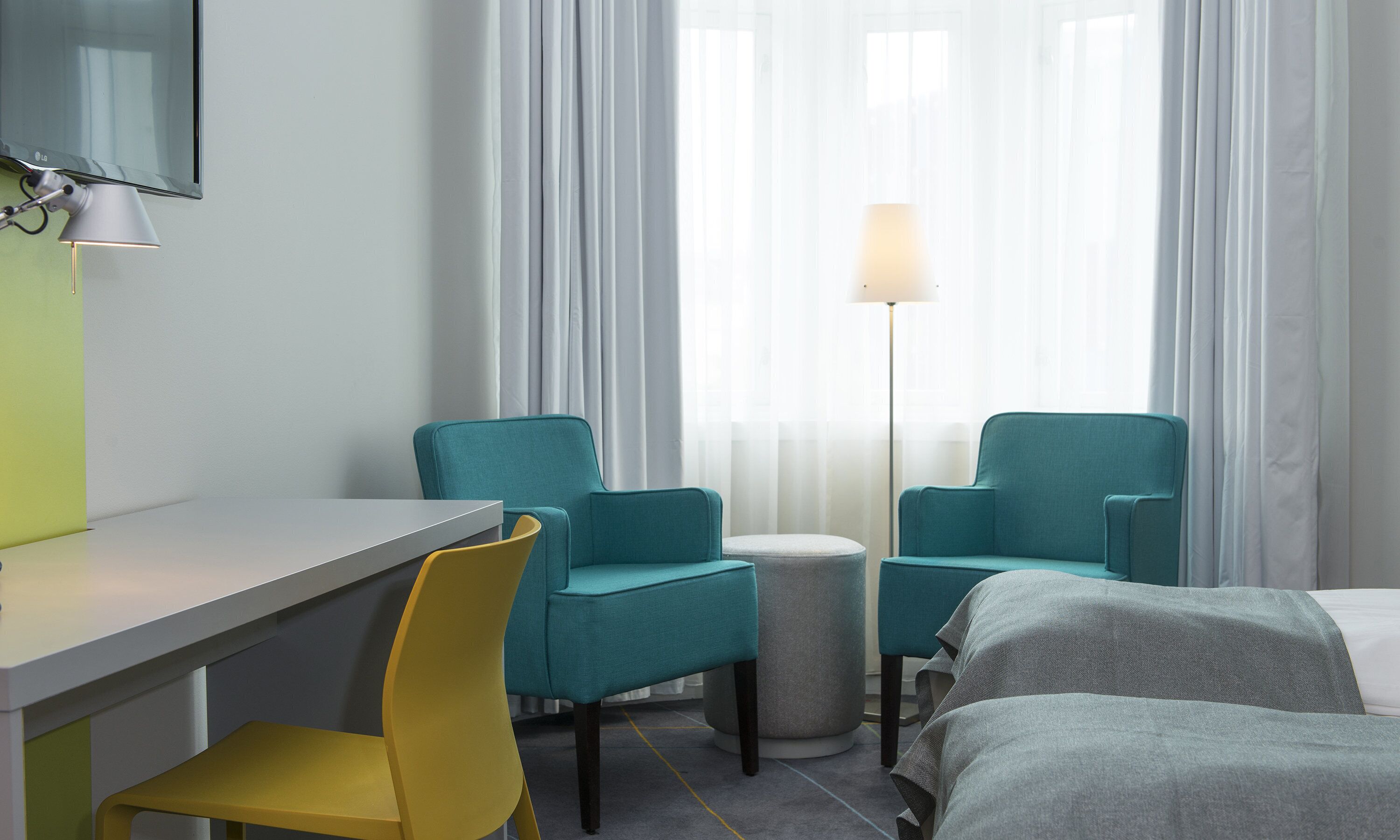 A hotel room with a desk, two teal chairs, a small round table, a lamp, and two beds.