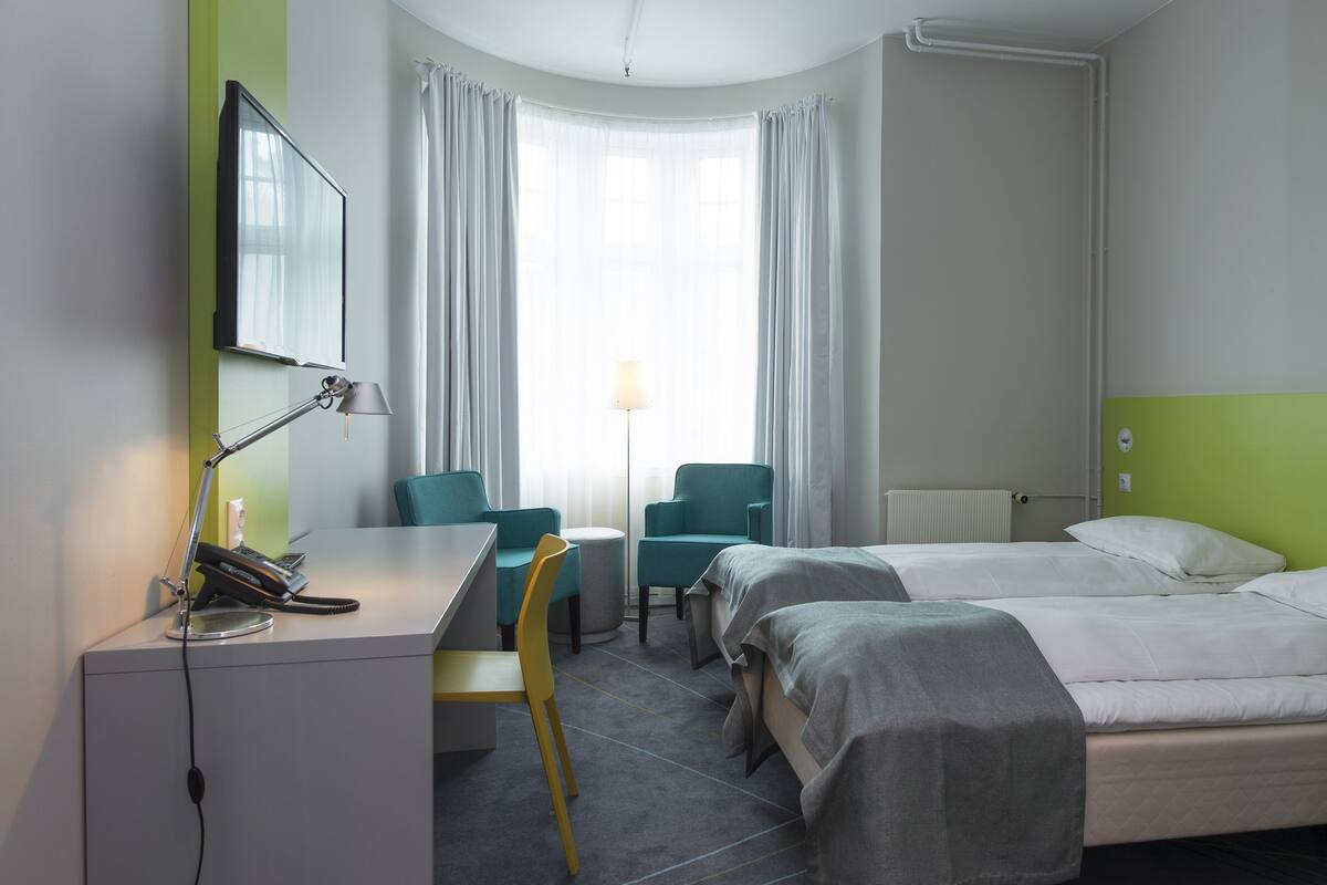 A hotel room with a desk, two chairs, a bed, and a window with curtains.